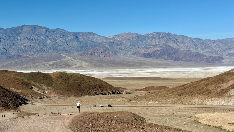 A person walking on a dirt road in a mountainous desert landscape with a vast valley and snow-like salt flats in the background under a clear blue sky.