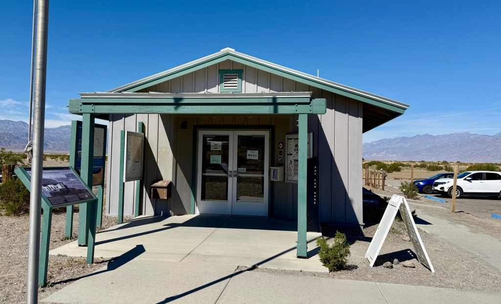 A small visitor center building in a desert landscape, featuring a covered entrance with glass doors, informational signage, and a sidewalk leading up to it.