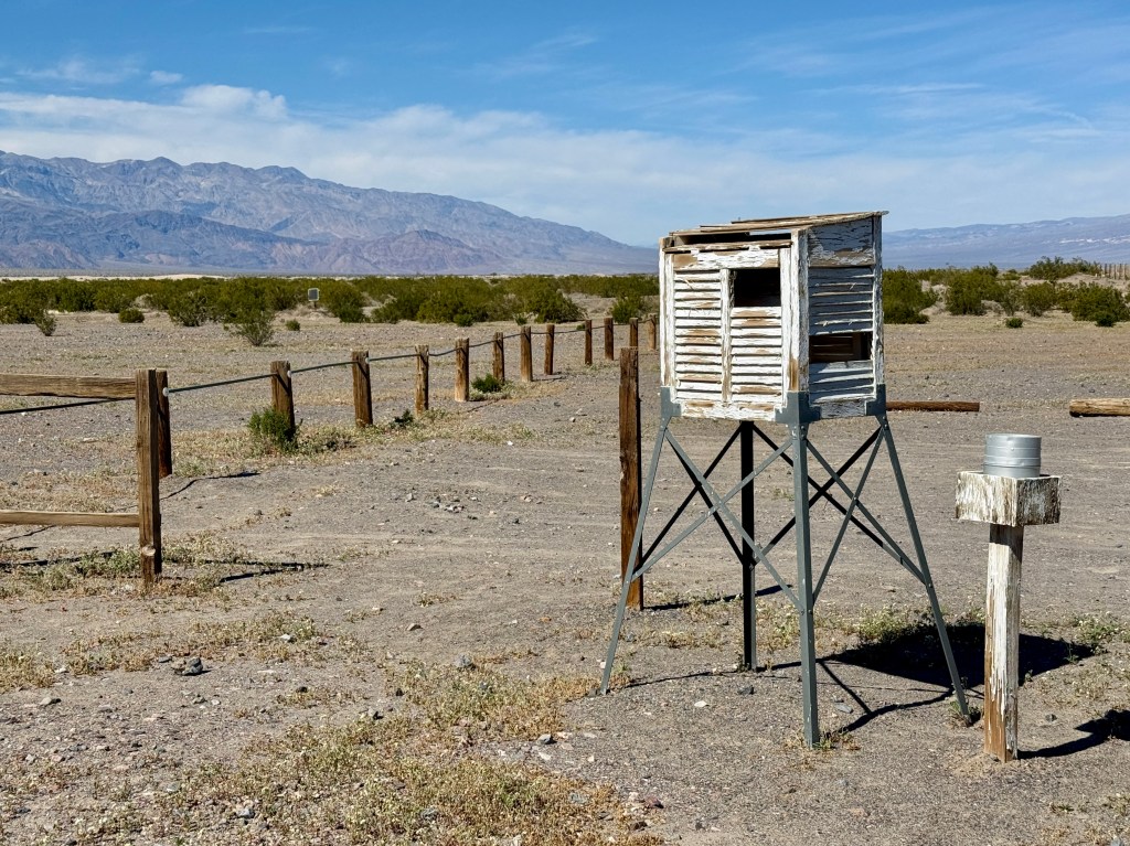 A weathered wooden observation tower on stilts in a barren desert landscape, with distant mountains and scattered shrubs in the background.
