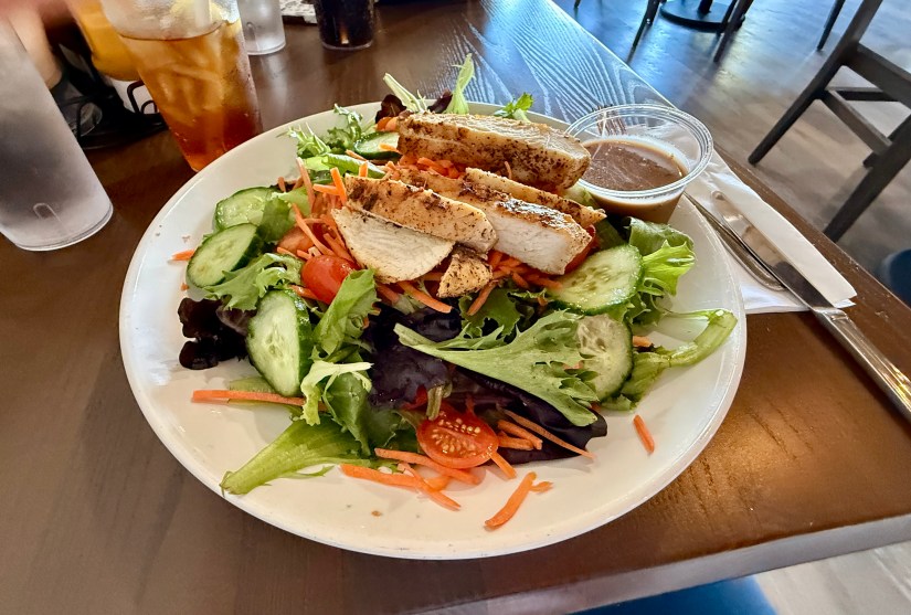 A plate of salad topped with grilled chicken breast slices, featuring lettuce, cucumbers, tomatoes, and shredded carrots, accompanied by a small container of dressing.