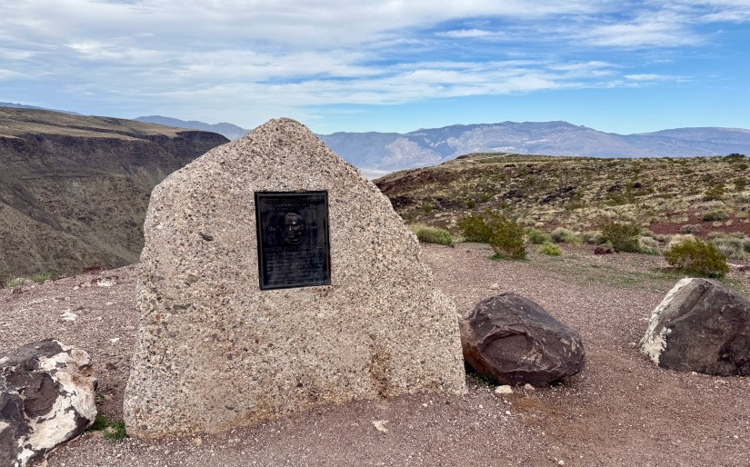 A prominent stone monument with a bronze plaque on a rocky landscape overlooking mountains and blue sky.