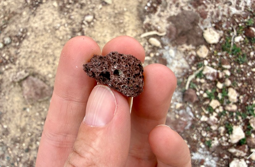 A person holding a piece of volcanic rock, showcasing its porous texture and brown color against a sandy and rocky background.