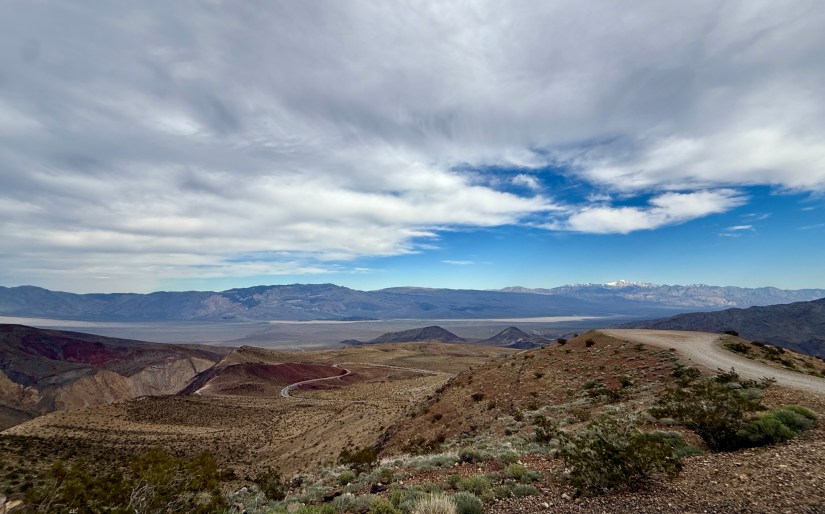 Scenic view of rugged mountains and valleys under a partly cloudy sky, with a winding dirt road visible in the foreground.
