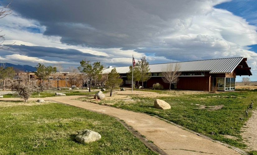 A view of a modern building with a metal roof set amidst green grass and scattered rocks, with a pathway leading to the entrance and trees around; a flagpole stands on the property, and dark clouds dominate the sky.