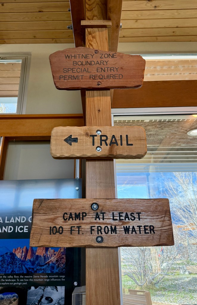 Wooden directional sign indicating Whitney Zone boundary with permit requirements, a trail to the left, and advice to camp at least 100 feet from water.