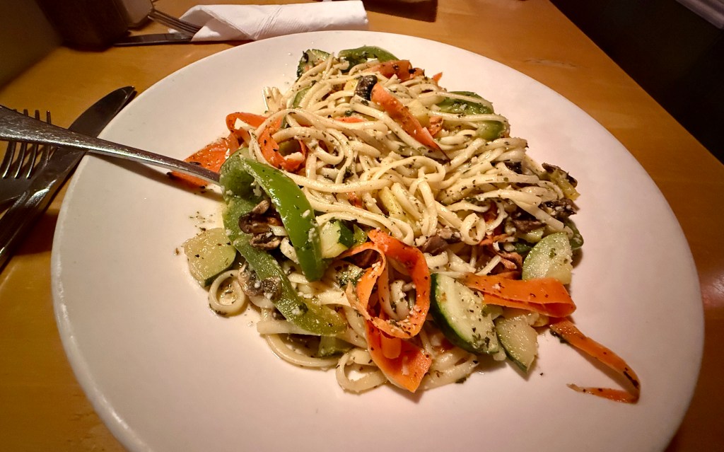 A plate of linguine pasta mixed with colorful sliced vegetables including green bell peppers, carrots, and zucchini, garnished with herbs.