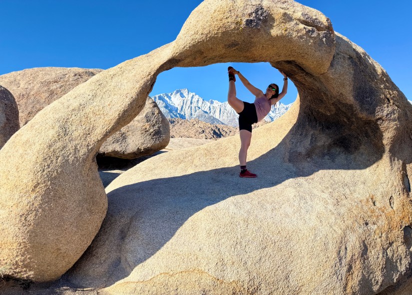 A person stretching one leg while standing under a large rock arch in a desert landscape with mountains in the background and a clear blue sky.