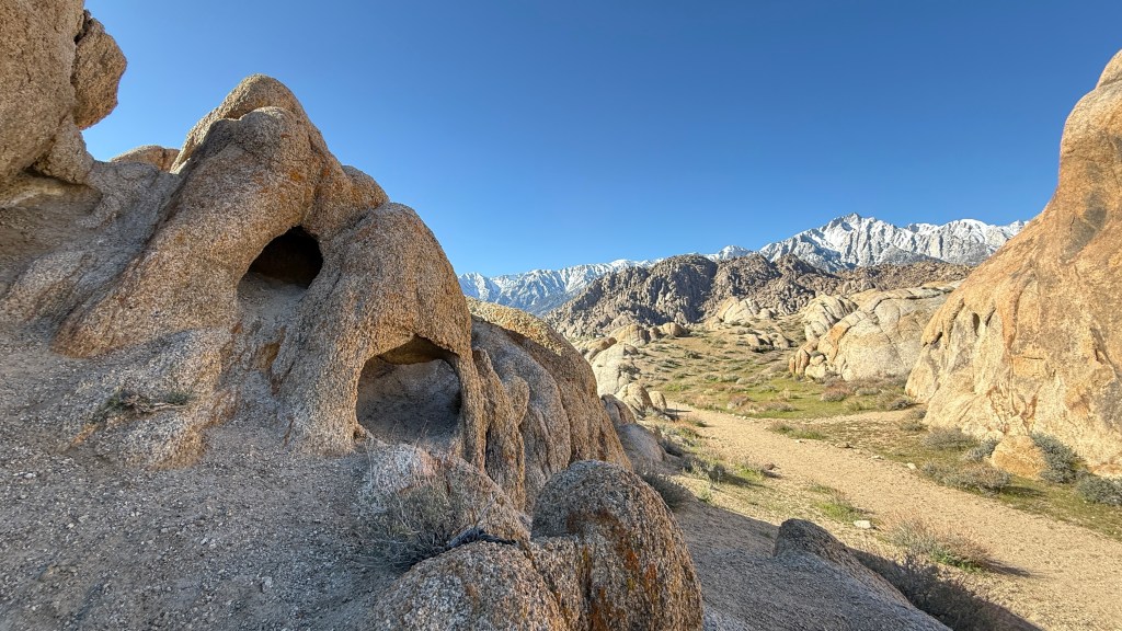 A rocky landscape featuring unique rock formations and a clear blue sky, with snow-capped mountains in the background.