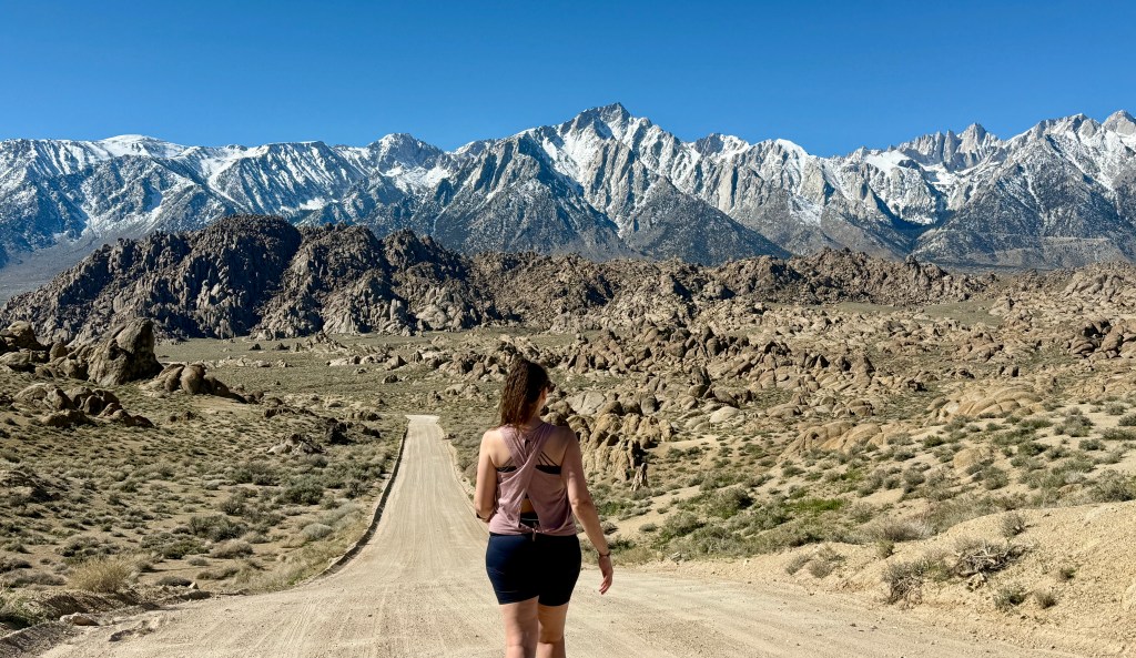 A woman walking on a dirt road surrounded by rocky terrain and snow-capped mountains under a clear blue sky.