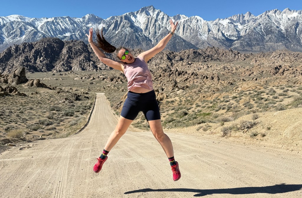 A woman jumping with joy on a dirt road surrounded by rocky terrain and snow-capped mountains in the background.