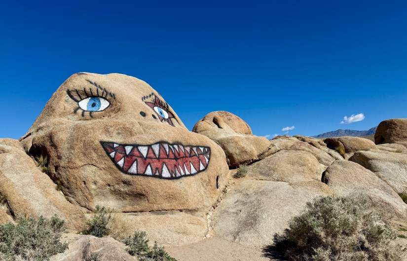 A large rock formation painted with a cartoonish face, featuring blue eyes and a wide, toothy grin, against a clear blue sky.