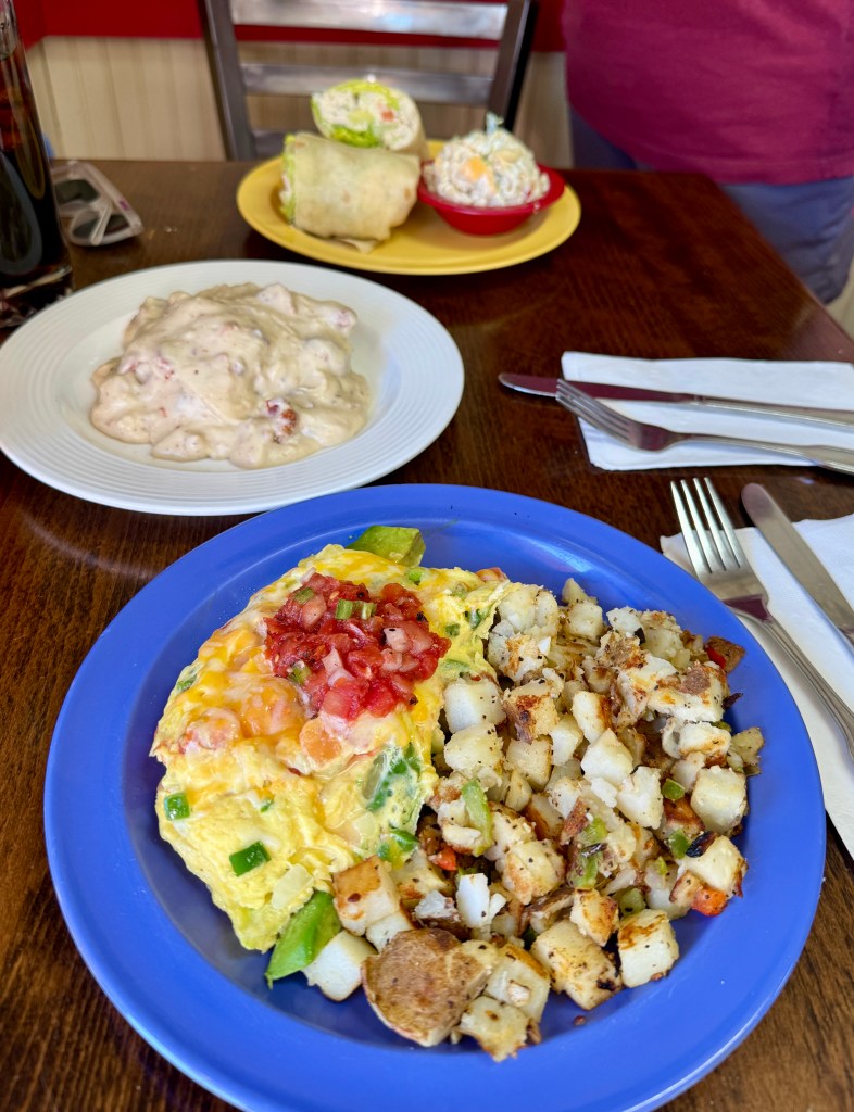A colorful breakfast plate featuring an omelette topped with tomatoes and green onions, served alongside golden-brown home fries. In the background, there are two additional dishes: a wrap and a bowl of salad.