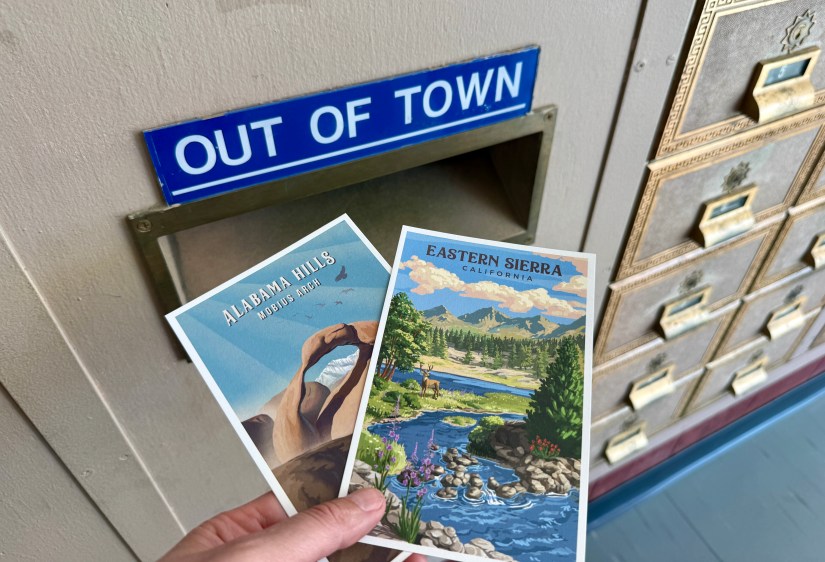 A hand holding two postcards, one featuring Alabama Hills and the other Eastern Sierra, near an 'OUT OF TOWN' mailbox.