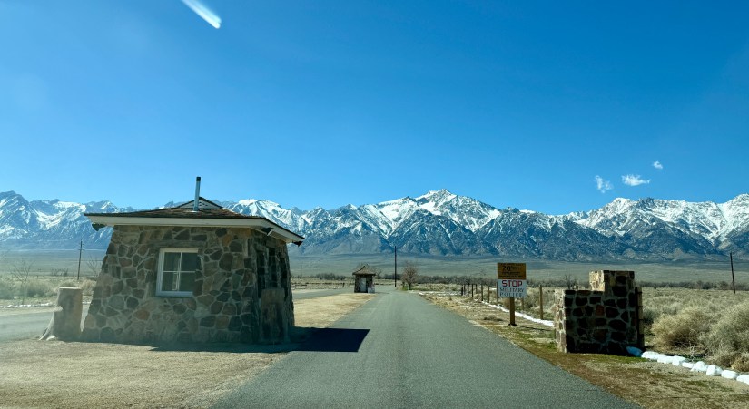 View of a stone entrance gate with a small building, leading to a road surrounded by snow-capped mountains under a clear blue sky.