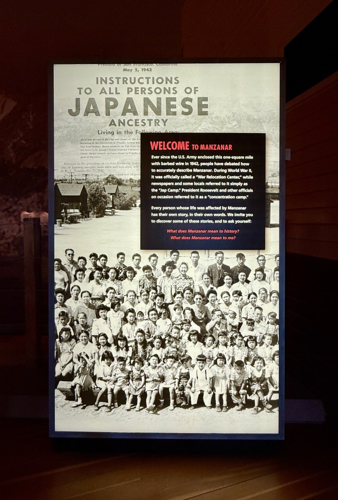A historical display at Manzanar featuring a large black and white photograph of a group of Japanese Americans, with instructions for individuals of Japanese ancestry and information about the internment during World War II.
