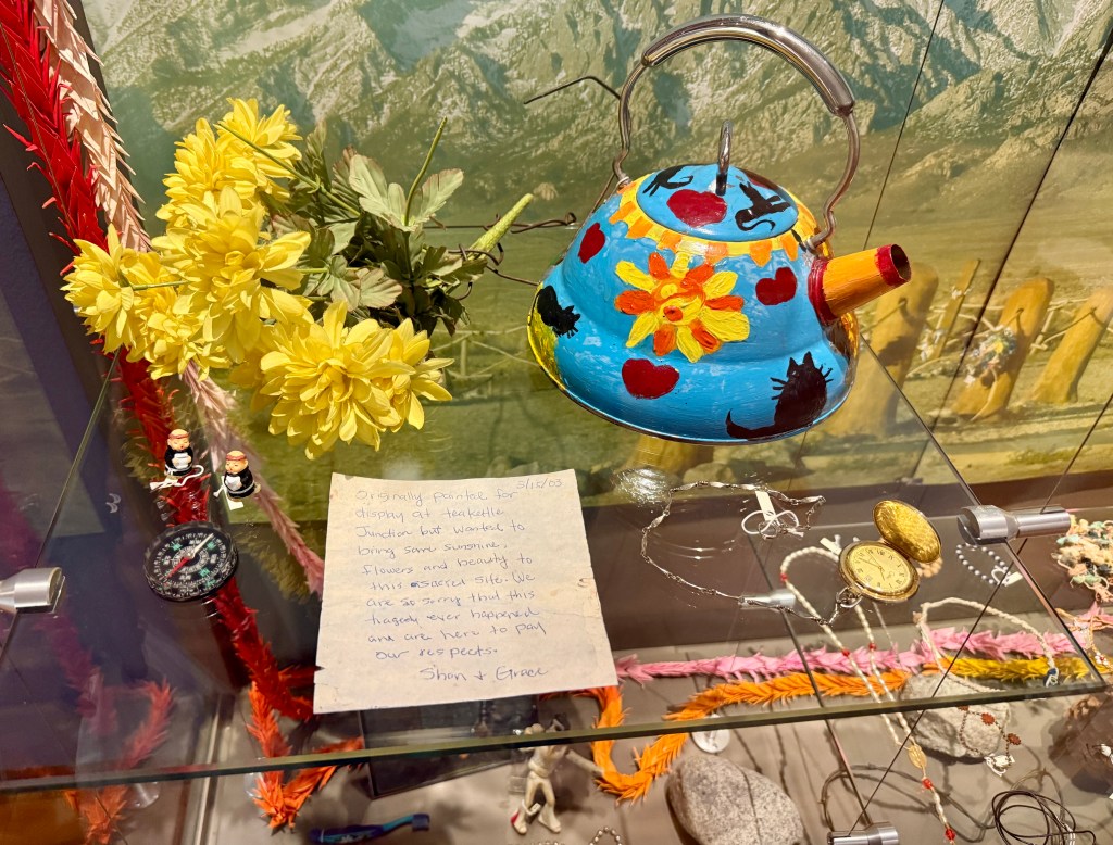 A colorful, hand-painted kettle decorated with flowers and hearts, placed among various decorative items and a handwritten note in a display case.