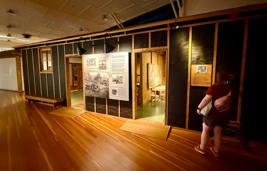 An individual explores an exhibition space featuring a wooden structure with an informative panel about gardens. The interior is visible through an open doorway, showing tables and displays.