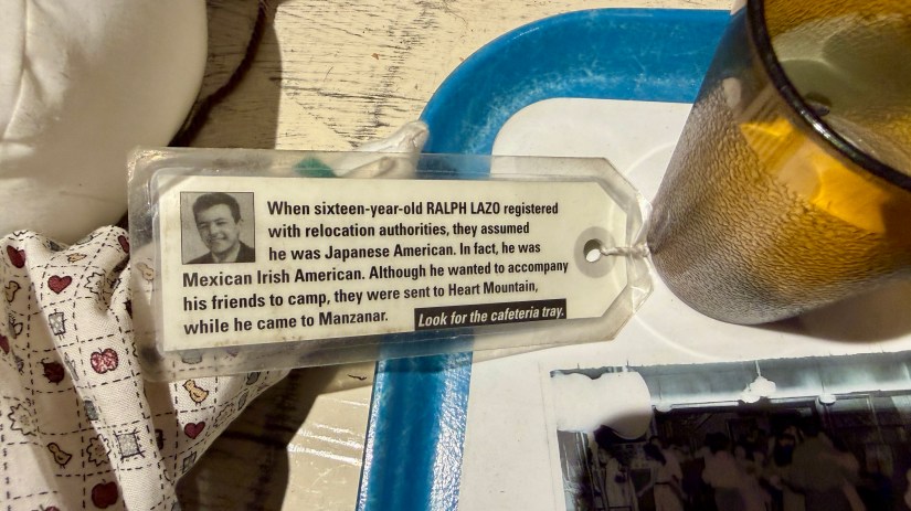 A close-up of a tag with a text about Ralph Lazo, a sixteen-year-old who registered with relocation authorities. The text discusses assumptions about his background and his desire to accompany friends to camp. A glass and tray are visible in the background.