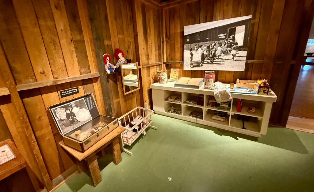 An interior view of a museum exhibit featuring wooden walls, a black and white photograph of a group of people outside, and various historical artifacts displayed on a shelf, including toys and books.