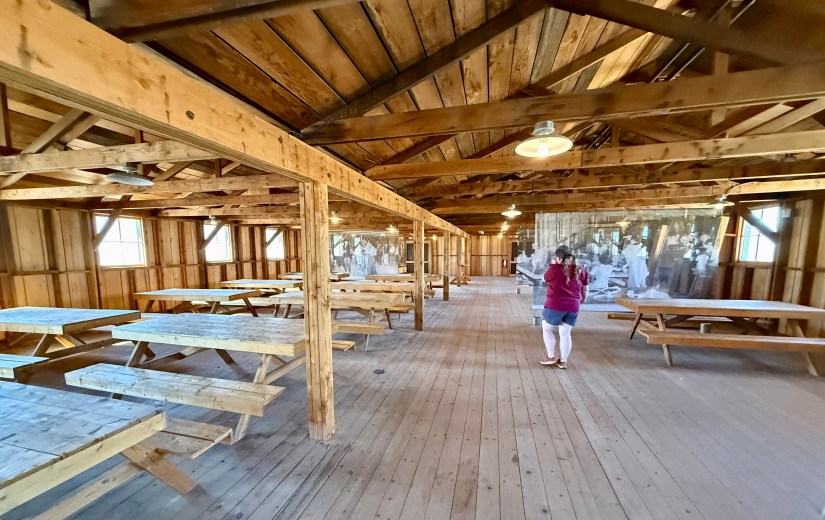 Interior of a wooden hall with picnic-style tables and a person taking a photo of a historical display.