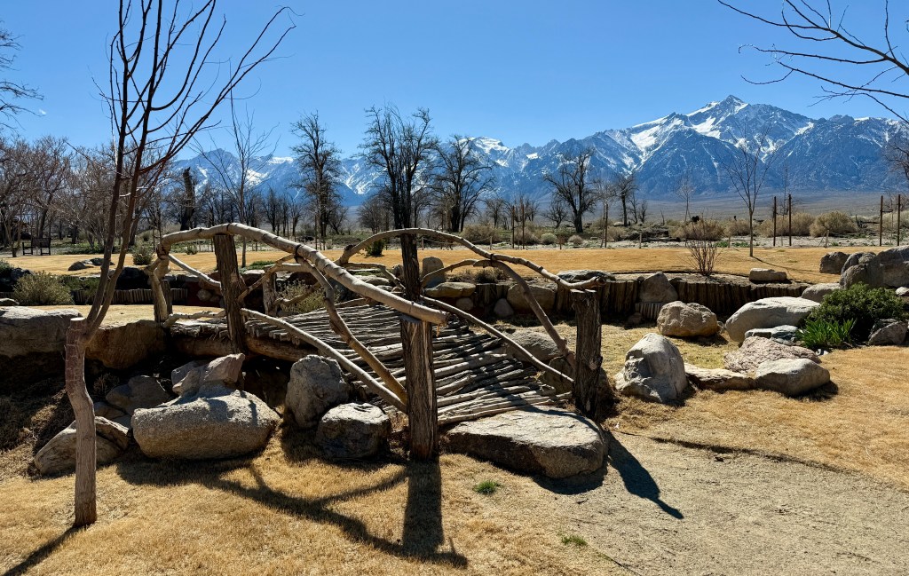 A wooden footbridge crossing over rocks and grass, with a backdrop of snow-capped mountains under a clear blue sky.