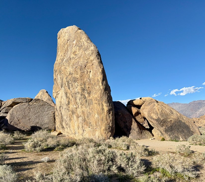 A tall, narrow rock formation stands amidst boulders and sparse vegetation under a clear blue sky.