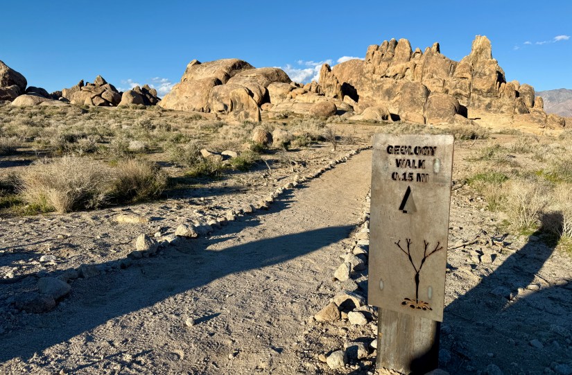 A sign for a geology walk indicating a distance of 0.15 miles, surrounded by rocky terrain and sparse vegetation under a clear blue sky.