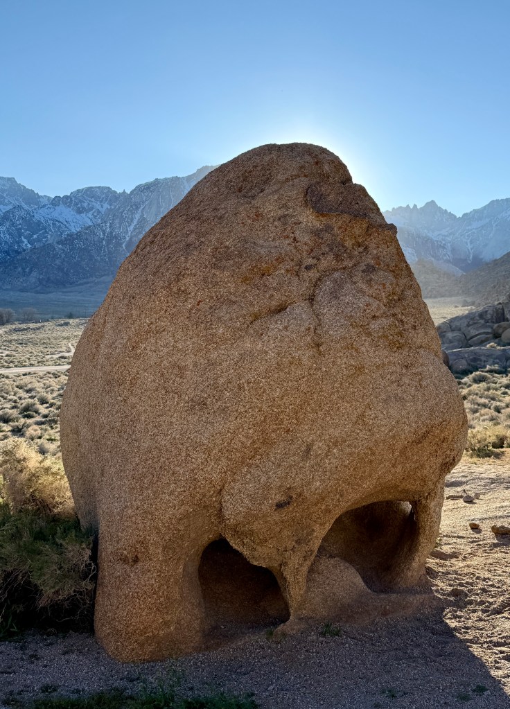 A large, rounded boulder with natural openings at its base, set against a backdrop of snow-capped mountains and a clear blue sky.