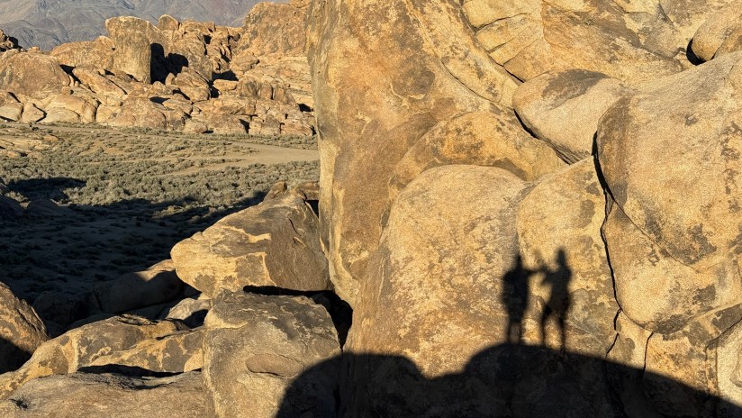 Shadows of two people standing on rocky terrain during sunset, surrounded by large boulders and sparse vegetation.