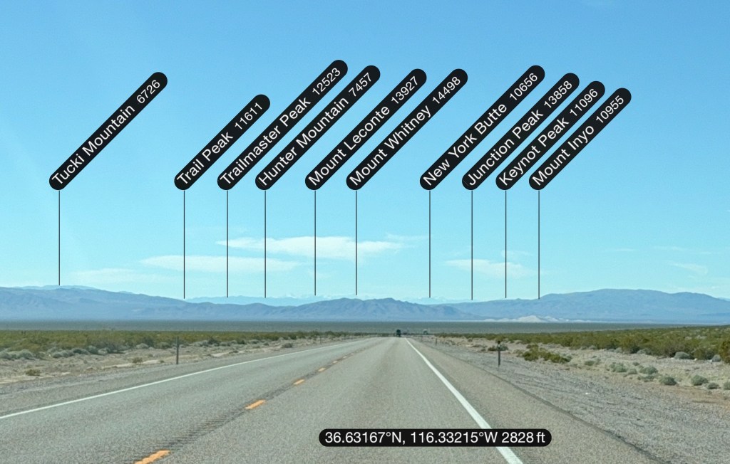 A road view in a desert with various mountain peaks labeled, showing their names and elevations, under a clear blue sky.