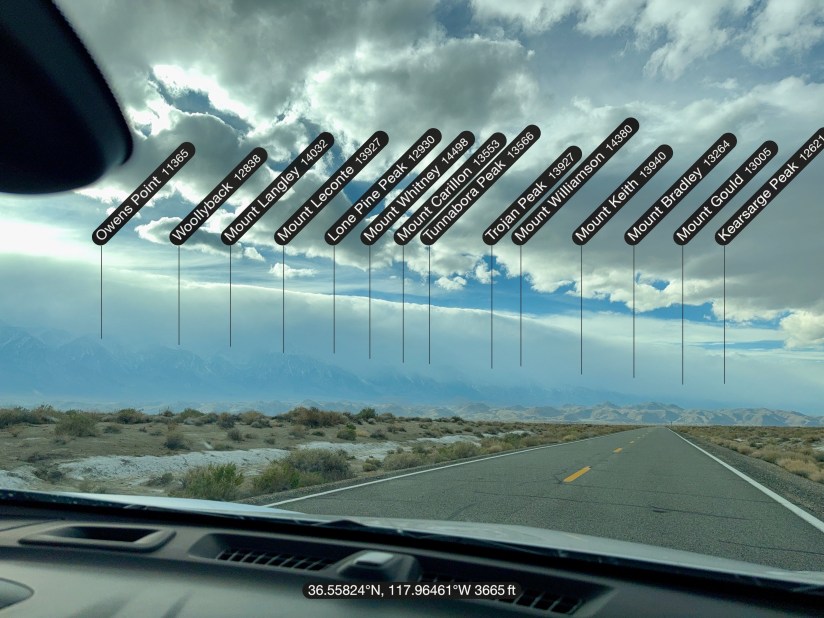 A view of a highway with mountains labeled in the distance, showing names and elevations of various peaks.