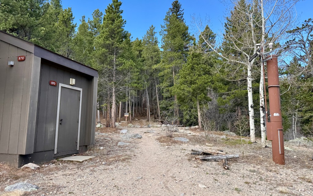 A dirt path leading through a forested area with dense green pine trees, alongside a utility shed marked with the number 870 and a metal pole on the right.
