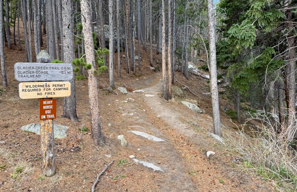 Sign at a trailhead indicating Glacier Creek Trail and Glacier Gorge with distance markers, surrounded by tall trees in a forest setting.