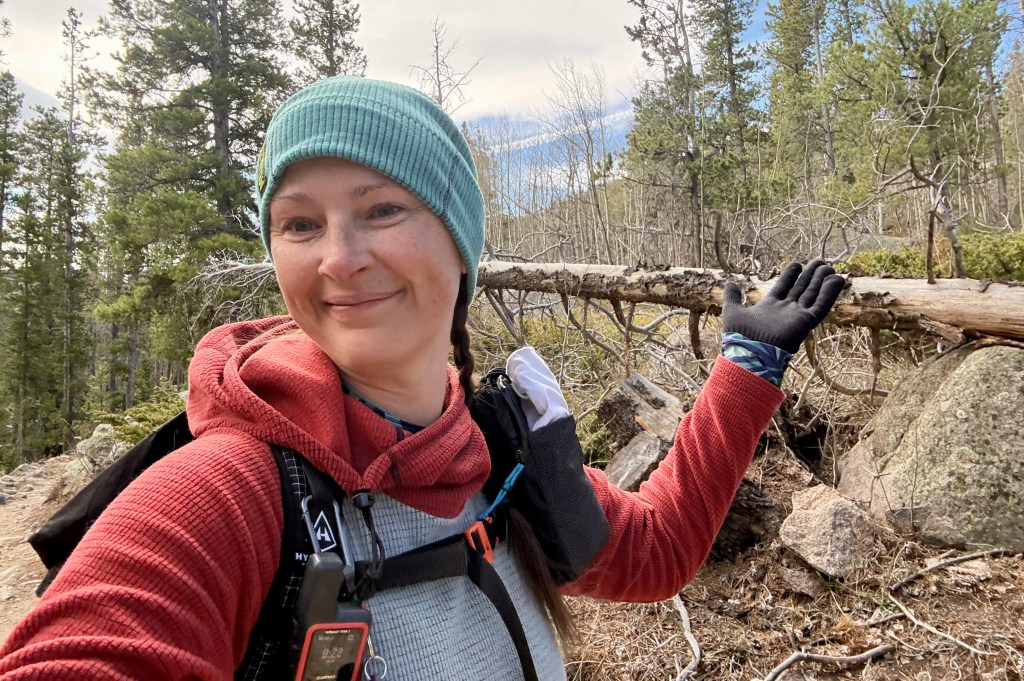 A smiling woman outdoors in a forest, wearing a light blue beanie and a red hoodie, posing with her left hand raised next to a fallen tree trunk, surrounded by greenery and rocks.