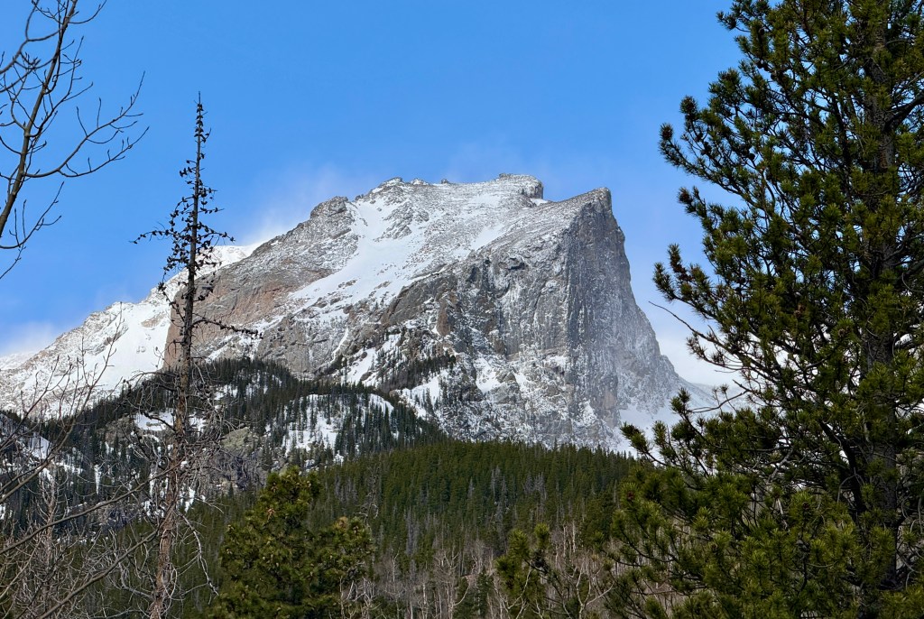 A snow-capped mountain peak with rocky cliffs, surrounded by lush green trees and a clear blue sky.