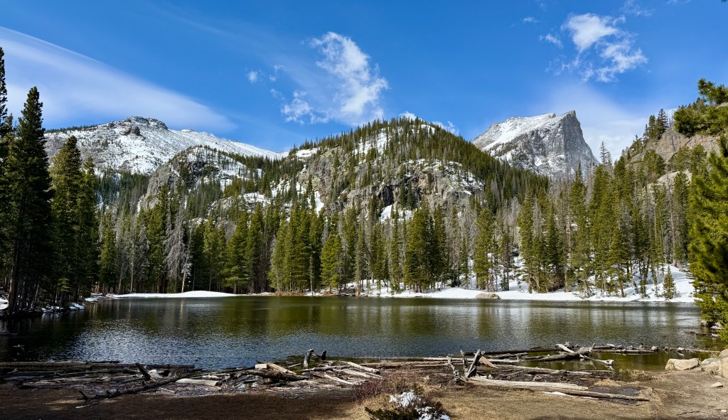 A serene mountain lake surrounded by tall evergreen trees and snow-capped peaks under a clear blue sky.