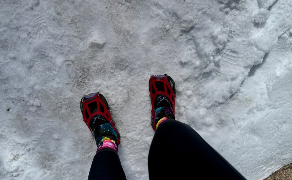 Close-up view of a person's feet wearing red crampons on snowy ground.