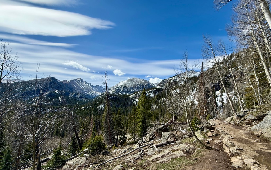 A scenic mountain landscape featuring a narrow hiking trail surrounded by evergreen trees and rocky terrain, with snow-capped mountains under a blue sky with wispy clouds.