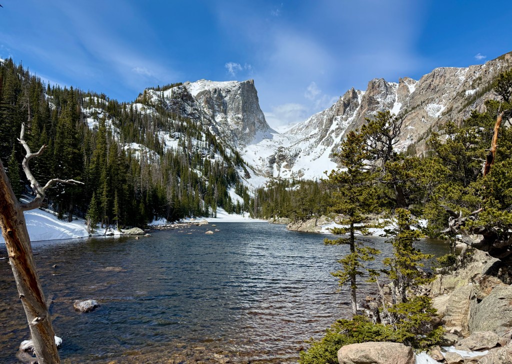 Scenic view of a mountain lake surrounded by snow-capped peaks and pine trees under a blue sky.