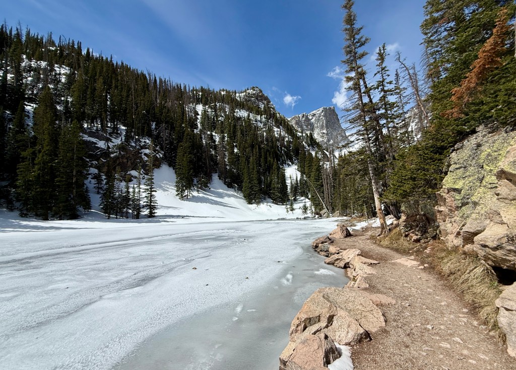 A scenic view of a frozen lake surrounded by snow-covered mountains and tall evergreen trees under a clear blue sky.