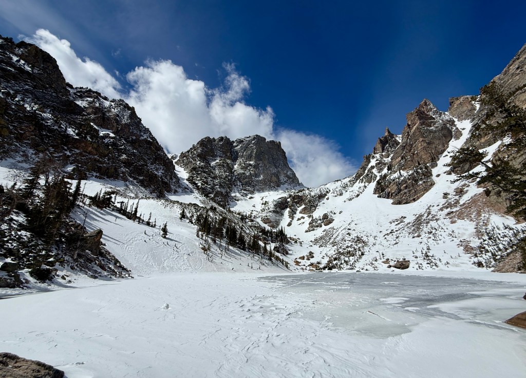 A scenic view of a snowy mountain landscape with towering rocky peaks under a bright blue sky and scattered clouds. A frozen lake is visible in the foreground, surrounded by trees and rugged terrain.