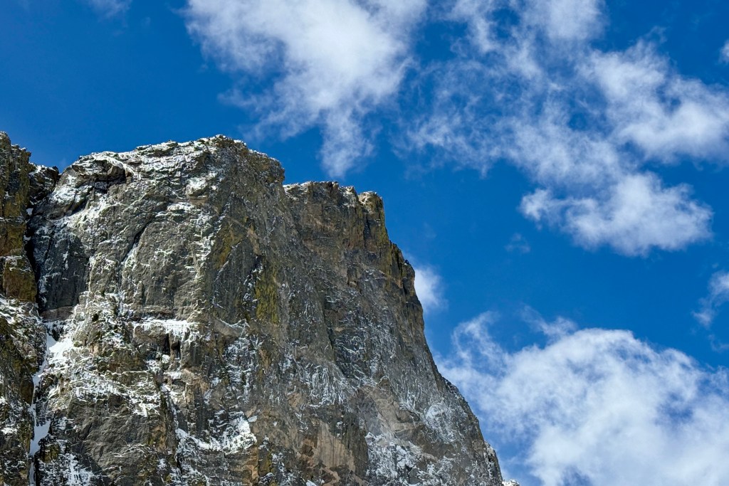 A close-up view of a rugged mountain peak covered with snow against a bright blue sky with fluffy white clouds.
