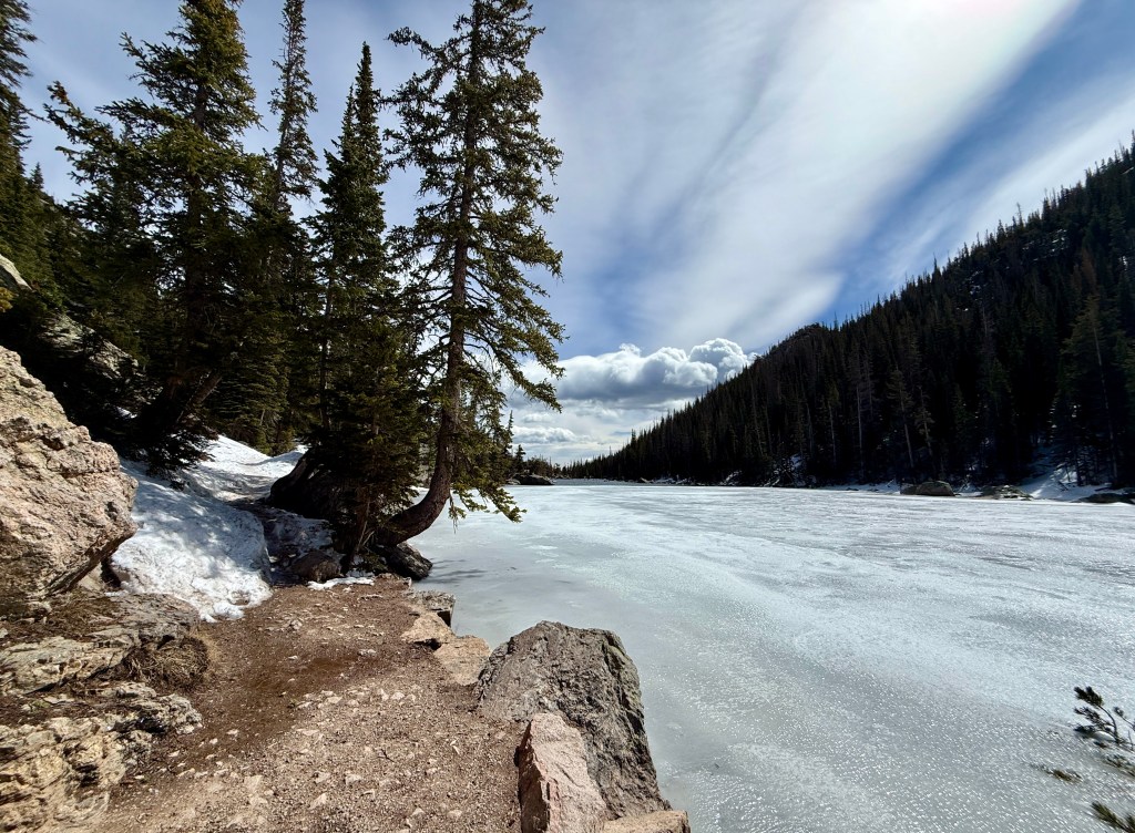 A serene winter landscape featuring a partially frozen lake surrounded by evergreen trees and mountains under a cloudy sky.
