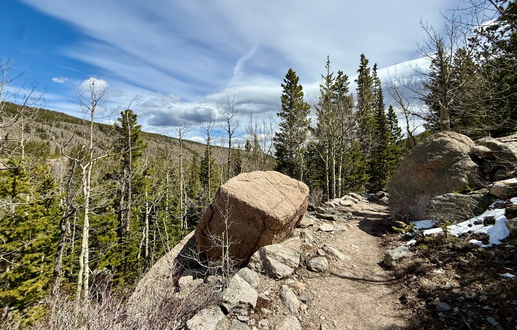 Rocky hiking trail surrounded by pine trees and boulders, under a partly cloudy sky.