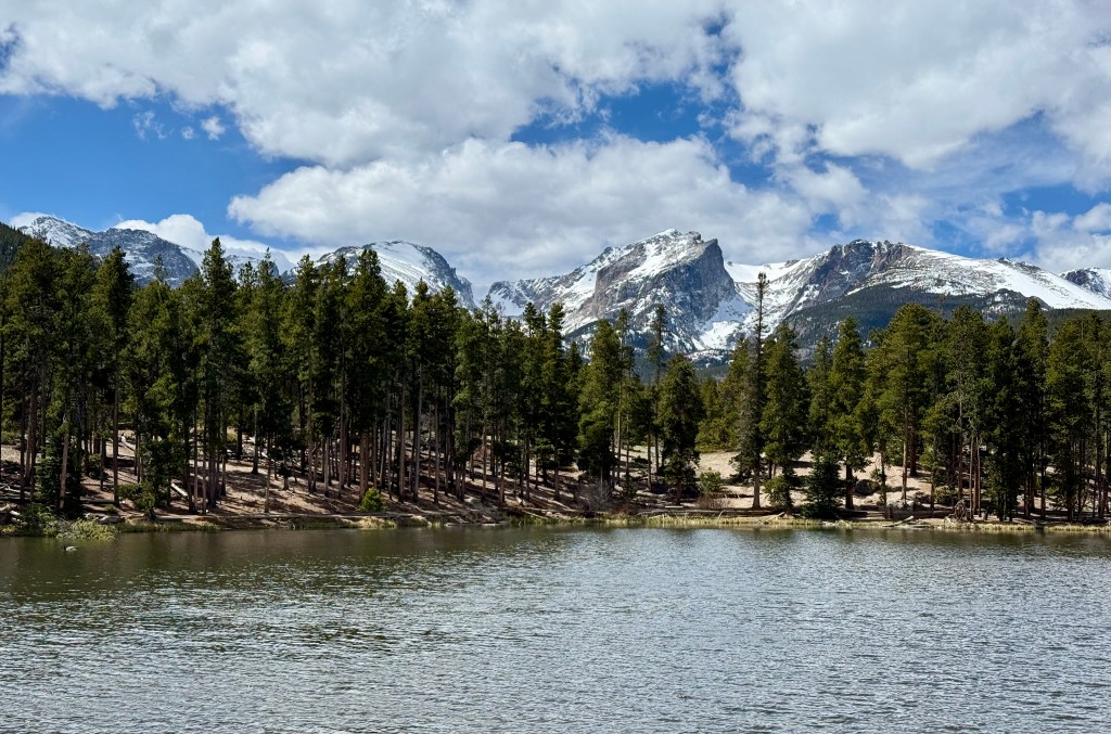Scenic view of a serene lake surrounded by tall pine trees and snow-capped mountains under a partly cloudy sky.