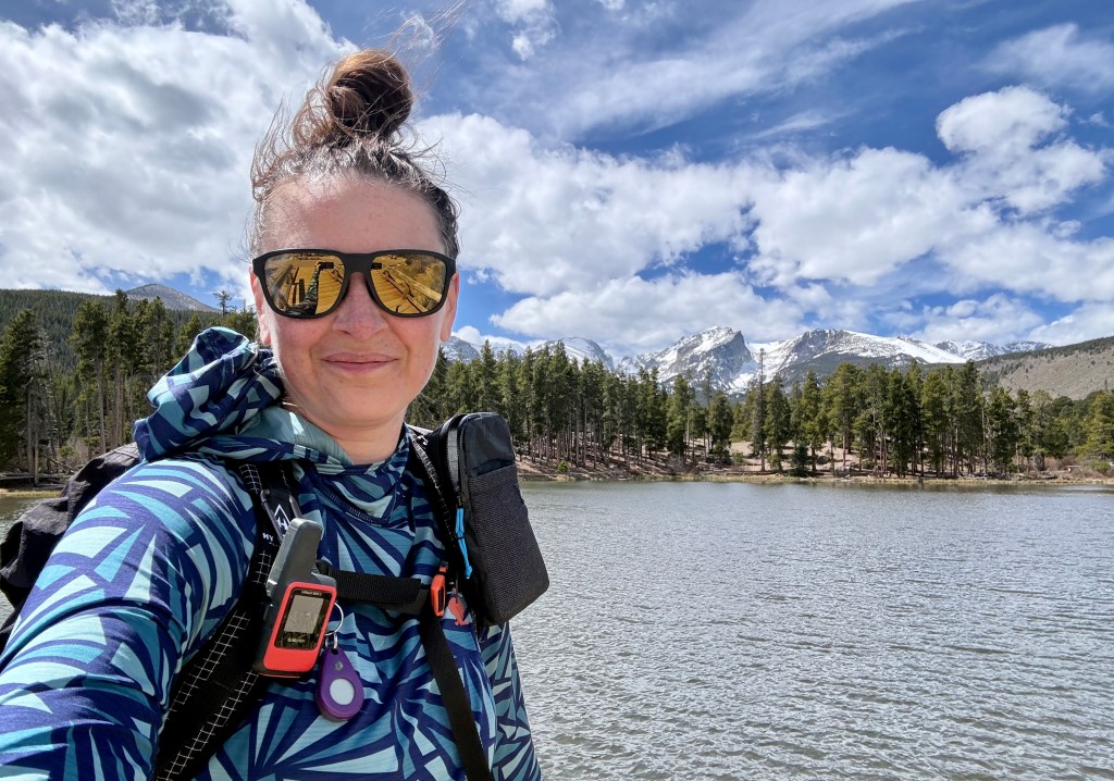 A person wearing sunglasses and a patterned jacket smiles for a selfie by a lake, surrounded by trees and mountains under a partly cloudy sky.