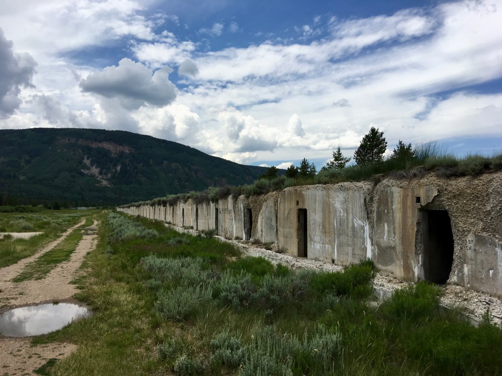 Remains of abandoned concrete structures along a dirt path in a mountainous landscape under a partly cloudy sky.
