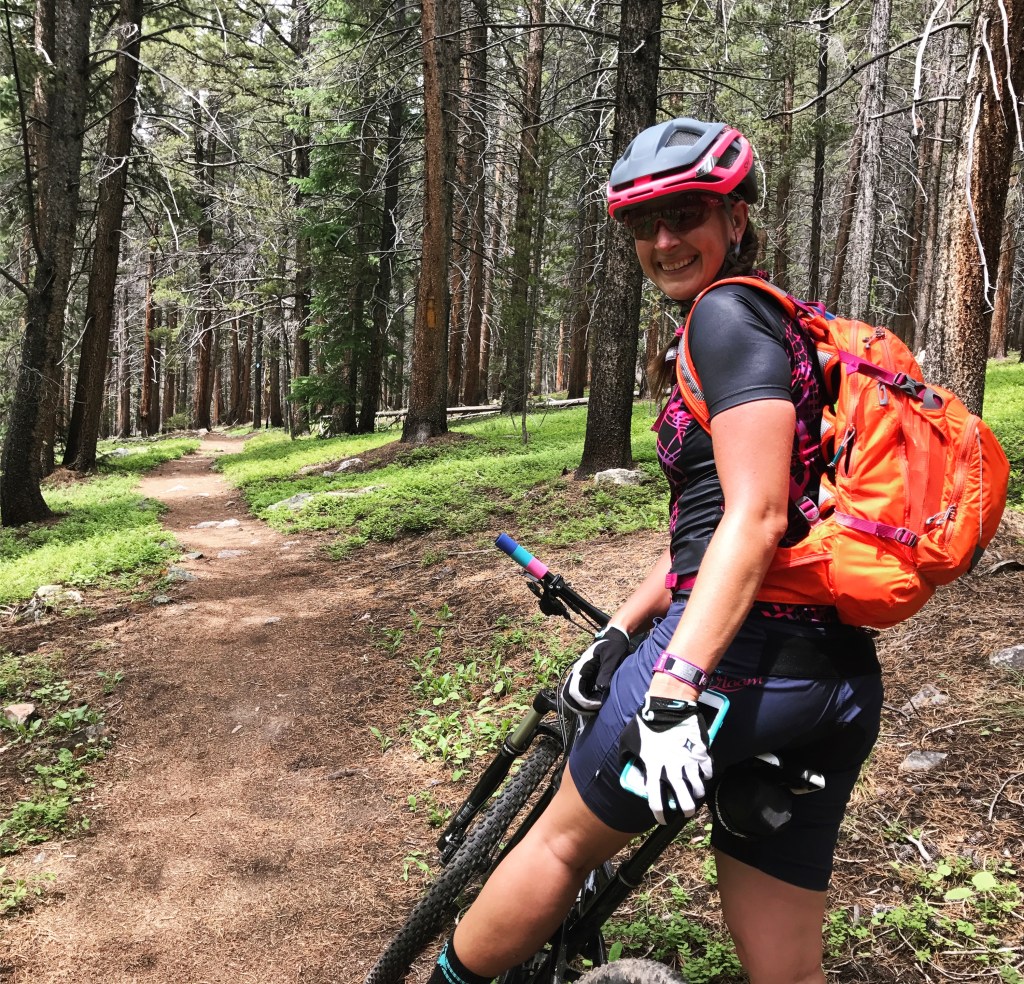 A female mountain biker resting with her bike on a dirt trail surrounded by trees and greenery.
