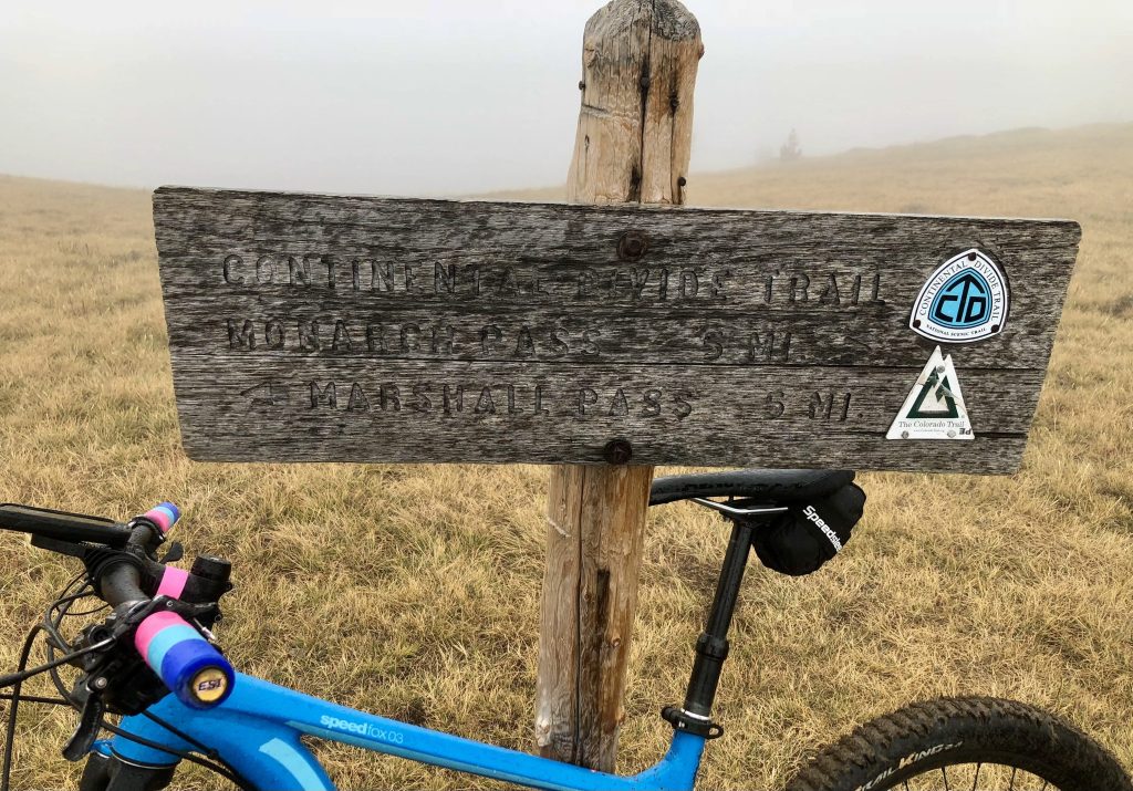 A wooden trail sign indicating directions and distances to the Continental Divide Trail and Monarch Pass, with a mountain bike parked beside it.