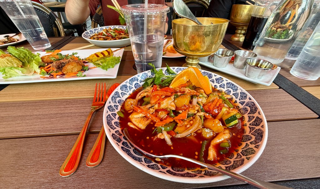 A colorful plate of seafood stir-fry with vegetables, garnished with an orange slice, served on a patterned dish, with additional dishes in the background.